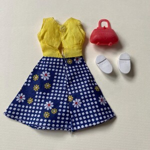 May include: A doll-sized outfit featuring a yellow vest and a blue skirt with white polka dots and daisy patterns. The set includes a red purse and white shoes, perfect for imaginative play.
