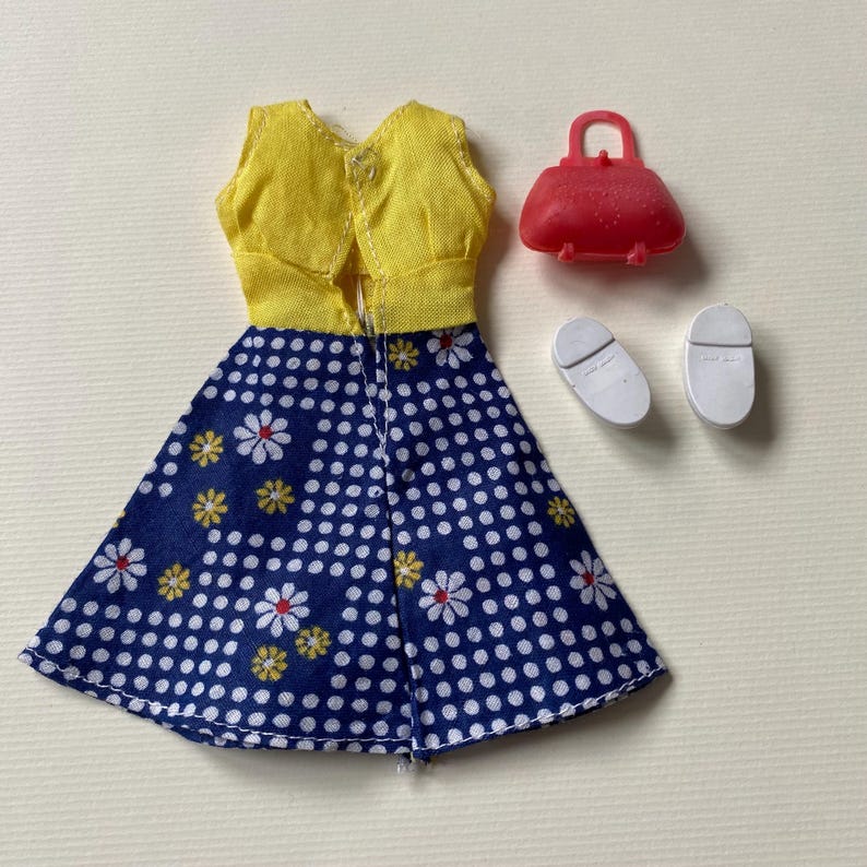 May include: A doll-sized outfit featuring a yellow vest and a blue skirt with white polka dots and daisy patterns. The set includes a red purse and white shoes, perfect for imaginative play.