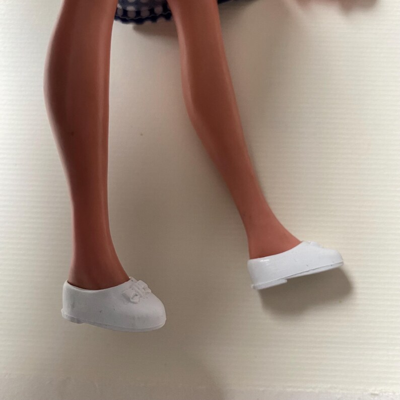 May include: A pair of white doll shoes with a bow detail. The shoes are a slip-on style and appear to be made of plastic. The doll's legs are visible above the shoes.