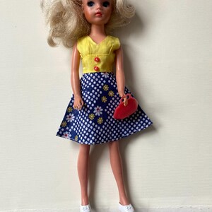 May include: A vintage doll with blonde hair, wearing a yellow top with red buttons and a blue polka dot skirt with white and yellow floral accents. The doll is holding a red purse and wearing white shoes.