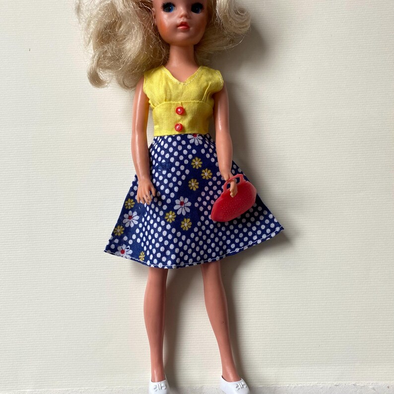May include: A vintage doll with blonde hair, wearing a yellow top with red buttons and a blue polka dot skirt with white and yellow floral accents. The doll is holding a red purse and wearing white shoes.