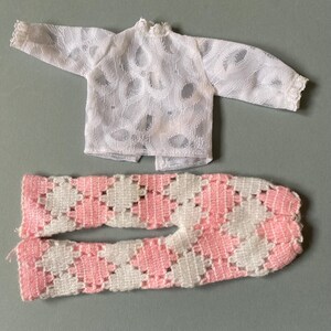 May include: A miniature outfit comprising a white, long-sleeved top and patterned pink and white trousers. The top has a high collar, and the trousers feature a diamond design. The outfit is crafted from a delicate, lace-like fabric.