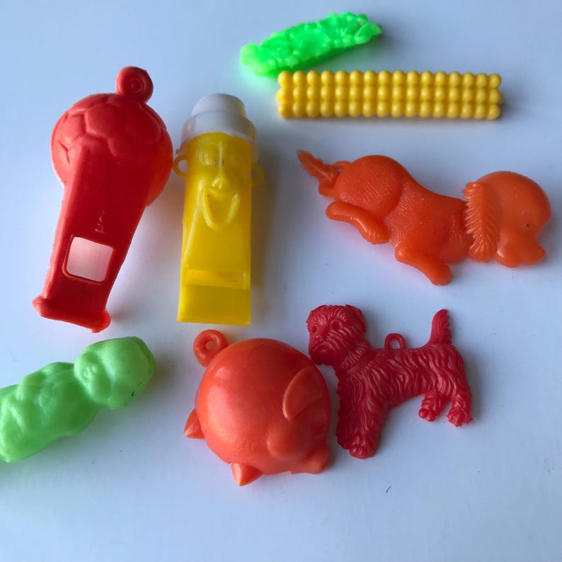 May include: A collection of colorful plastic toys, including a whistle shaped like a face, a whistle shaped like a soccer ball, a pig, a dog, a corn cob, and a green worm.