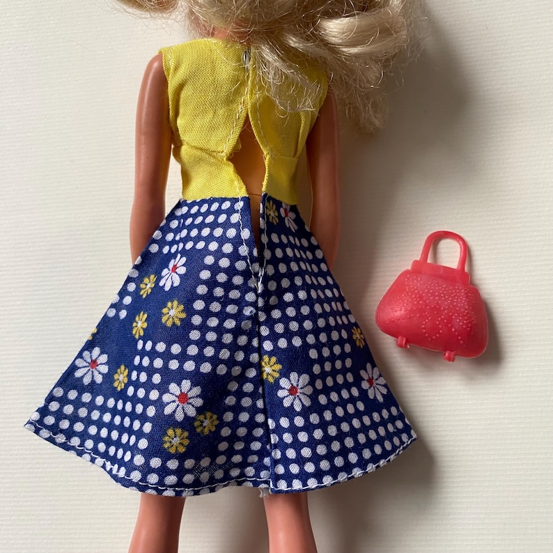 May include: A doll wearing a yellow top and a blue skirt with white polka dots and floral accents. A small red purse with a handle sits next to the doll. The doll's blonde hair is visible.