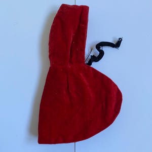 May include: A small, red velvet hooded cape with a rounded bottom. The cape has a hood and a black tie. The fabric appears soft and plush. The cape is likely for a doll or a costume.