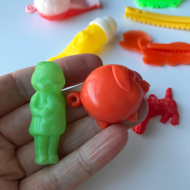May include: A set of vintage plastic toys, including a green girl doll, an orange pig, and a red dog. The toys are all small and have a retro look.