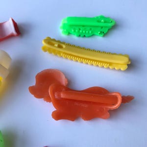 May include: A set of four colorful plastic hair clips. The clips are shaped like a dog, a flower, a saw, and a zig-zag. The clips are all in bright colors, including green, yellow, orange, and white.