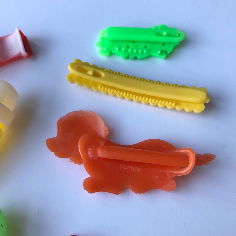 May include: A set of four colorful plastic hair clips. The clips are shaped like a dog, a flower, a saw, and a zig-zag. The clips are all in bright colors, including green, yellow, orange, and white.