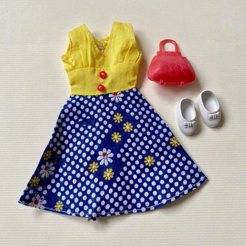 May include: A doll-sized outfit featuring a yellow top with red buttons and a blue polka-dot skirt with white and yellow floral accents. Includes a red purse and white shoes, evoking a vintage aesthetic.