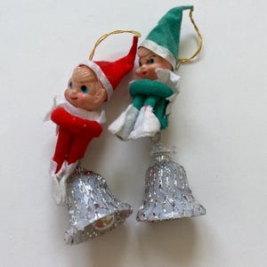 May include: Two hanging ornaments featuring small elf figures with painted faces. One elf wears a red suit and hat, the other a green suit and hat. Each elf is attached to a white bell covered in a silver metallic mesh netting with a gold string.