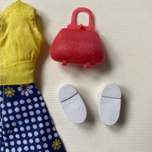 May include: A doll's outfit featuring a yellow top, a blue skirt with white polka dots, a red purse, and two white shoes. The outfit is made of plastic and fabric. The purse has a handle and the shoes have a curved shape.