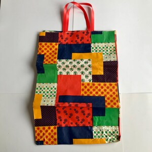 May include: A patchwork tote bag with a red ribbon handle. The bag is made up of various geometric patterns in red, yellow, green, blue, and white. The bag is rectangular and appears to be made of a durable material.