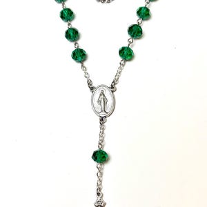 May include: A rosary necklace featuring faceted green beads and a silver-tone crucifix pendant. The necklace includes a central oval pendant with a religious figure. The beads are strung on a silver chain.