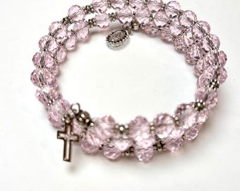 Könnte beinhalten: Ein Wickelarmband mit hellrosa facettierten Perlen und silberfarbenen Akzenten. Das Armband hat einen kleinen Kreuzanhänger und einen runden Anhänger. Die Perlen sind mit silberfarbenem Draht aufgefädelt.