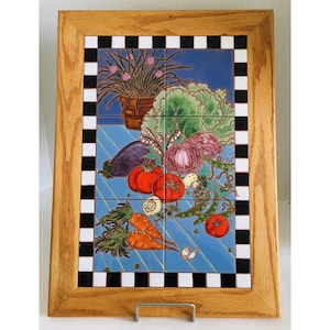May include: A framed ceramic tile art piece featuring a still life of vegetables. The artwork includes tomatoes, carrots, an eggplant, and other produce, set against a blue background. The frame is made of light-colored wood, with a black and white checkered border.