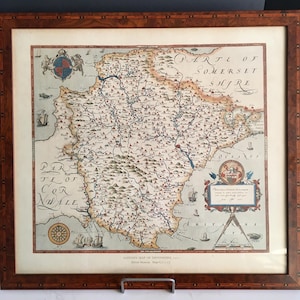 May include: A framed antique map of Devonshire, England, from 1571, with intricate details and vibrant colors. The map is housed in a dark wood frame, showcasing the historical cartography and adding a touch of vintage charm to the artwork.