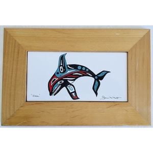 May include: A framed artwork featuring a stylized orca whale design. The orca is depicted in black, red, and blue against a white background. The artwork is in a light-colored wooden frame. The word "Orca" is written on the bottom left.