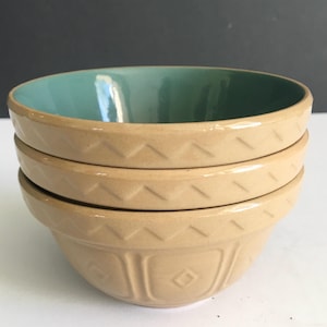May include: A stack of three beige ceramic mixing bowls. The bowls have a teal interior and a textured exterior with a repeating geometric pattern. The bowls are nested together, showcasing their varying sizes and design.