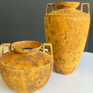 May include: Two mustard yellow vases with a weathered, textured finish. Both vessels feature a wide circular opening and two small, rectangular handles on opposite sides. The larger vase is tall and tapered, while the smaller is round.
