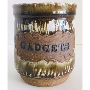 May include: A ceramic jar with a brown and white glaze. The word "GADGETS" is embossed in a brown, decorative label on the front. The jar has a cylindrical shape with a slightly flared base and a dark brown rim.