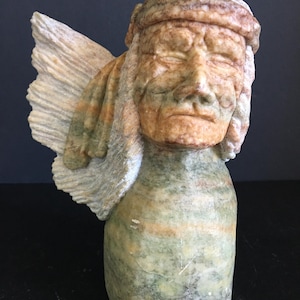 May include: A carved stone sculpture of a figure with closed eyes, wearing a head covering and a wing. The sculpture is primarily green, with brown and tan accents. The figure has a serene expression.