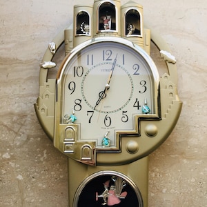 May include: Gold-colored quartz wall clock with a castle design. The clock face has black numerals and hands, with decorative figures and a pendulum. The clock has three small towers at the top and a decorative bottom.