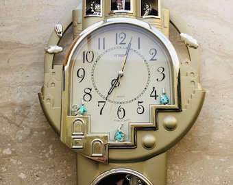 May include: Gold-colored quartz wall clock with a castle design. The clock face has black numerals and hands, with decorative figures and a pendulum. The clock has three small towers at the top and a decorative bottom.