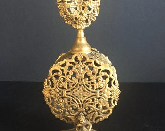 May include: Ornate gold-colored perfume bottle with a filigree design. The bottle features a round base, a detailed body with a cherub, and a floral-shaped top. The intricate metalwork creates a vintage aesthetic.
