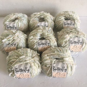 May include: Eight skeins of textured yarn in shades of green, white, and peach. Each skein has a label that reads "Butterfly" with a dog illustration. The yarn appears soft and fluffy, ideal for knitting or crochet projects.