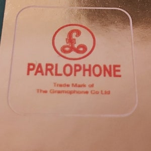 以下が含まれることがあります： 角が丸い透明な正方形のステッカー。赤い文字で「PARLOPHONE」と、スタイリッシュな「£」記号の赤い円形ロゴが特徴です。テキストの下には「Trade Mark of The Gramophone Co Ltd.」と書かれています。