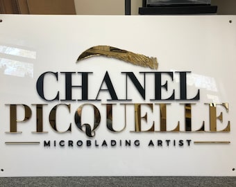 May include: White sign with black and gold lettering. The sign reads "Chanel Picquelle Microblading Artist". The sign has a gold eyebrow shape above the text.