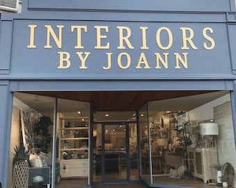 May include: A storefront with a blue facade and large gold lettering that reads "Interiors by Joann". The storefront has large windows that display a variety of home decor items, including furniture, lamps, and artwork.