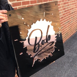 Op de afbeelding: Een zwart metalen bord met een koperkleurig splatterontwerp. Het bord zegt "Bob Photography" in een gestileerd lettertype.