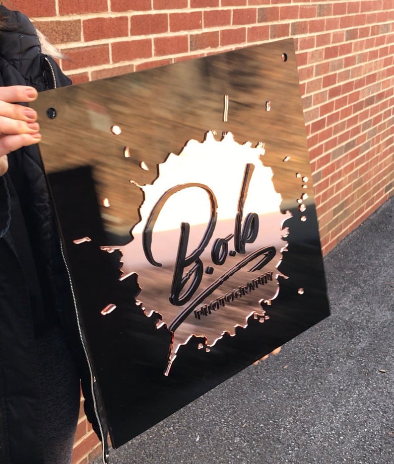 Op de afbeelding: Een zwart metalen bord met een koperkleurig splatterontwerp. Het bord zegt "Bob Photography" in een gestileerd lettertype.