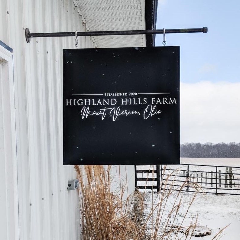 Puede incluir: Un letrero negro con texto blanco que dice "Establecido 2020 Highland Hills Farm Mount Vernon, Ohio". El letrero est&aacute; colgado de una barra de metal unida a un edificio blanco.