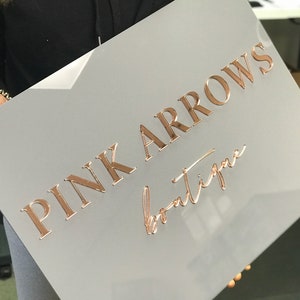 Op de afbeelding: Een grijs bord met de tekst "PINK ARROWS boutique" in roze gouden letters.