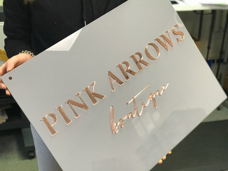Op de afbeelding: Een grijs bord met de tekst "PINK ARROWS boutique" in roze gouden letters.