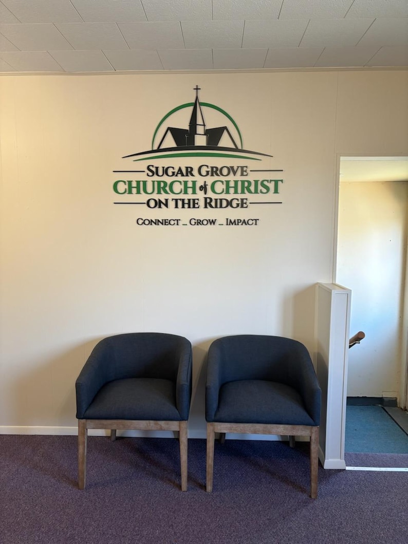 May include: Two dark gray upholstered chairs with light wooden legs positioned in front of a wall featuring the black and green logo and text for Sugar Grove Church of Christ on the Ridge, including the words Connect, Grow, and Impact.