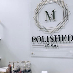 Op de afbeelding: Een gouden en zwart bord met de tekst "POLISHED BY MAI" en een gouden geometrisch frame met de letter "M" erin. Het bord is gemonteerd op een witte muur. Er staan ​​verschillende potten met kaarsen op een tafel voor het bord.