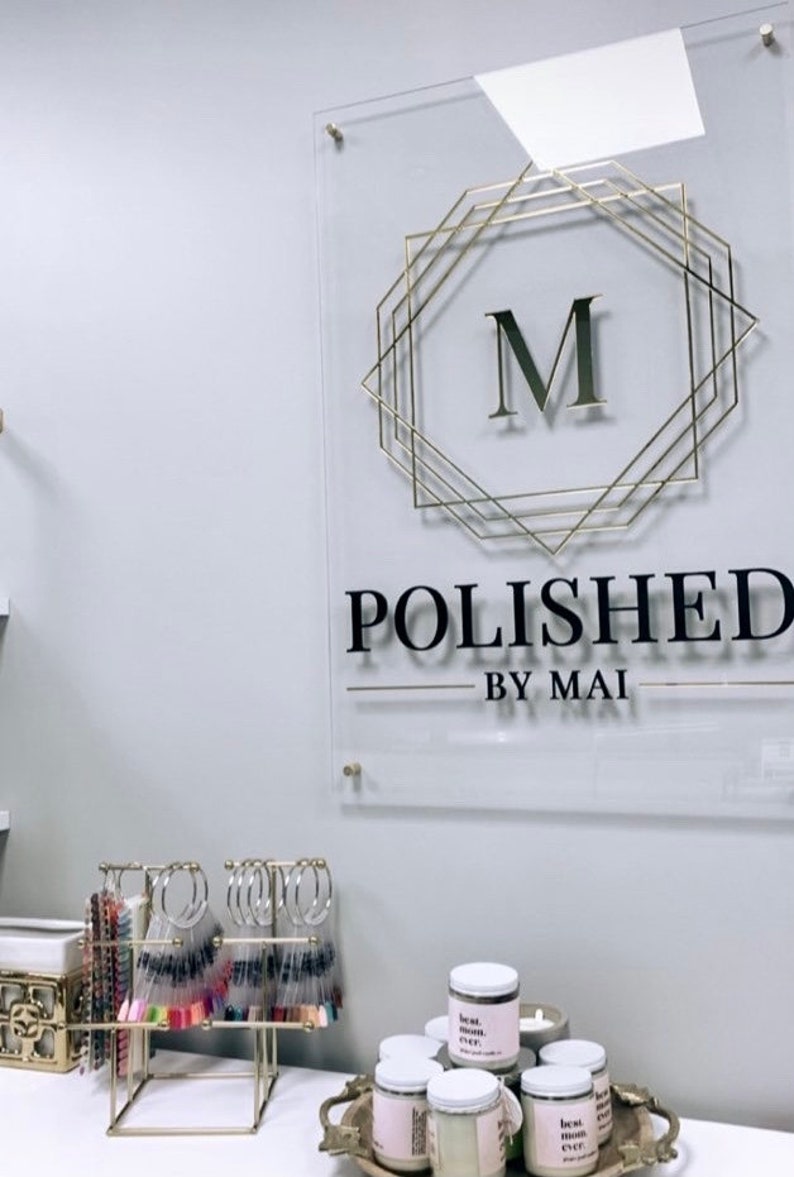 Op de afbeelding: Een gouden en zwart bord met de tekst "POLISHED BY MAI" en een gouden geometrisch frame met de letter "M" erin. Het bord is gemonteerd op een witte muur. Er staan ​​verschillende potten met kaarsen op een tafel voor het bord.