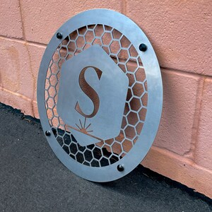 May include: A circular metal sign with a honeycomb pattern and a cutout of the letter "S" in a hexagon shape. The sign has a silver finish and is mounted on a pink wall.