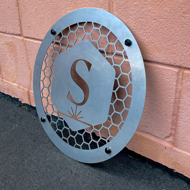 May include: A circular metal sign with a honeycomb pattern and a cutout of the letter "S" in a hexagon shape. The sign has a silver finish and is mounted on a pink wall.