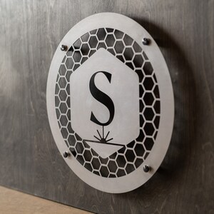 May include: A silver metal sign with a honeycomb pattern and the letter "S" in the center. The sign is mounted on a dark wood surface.