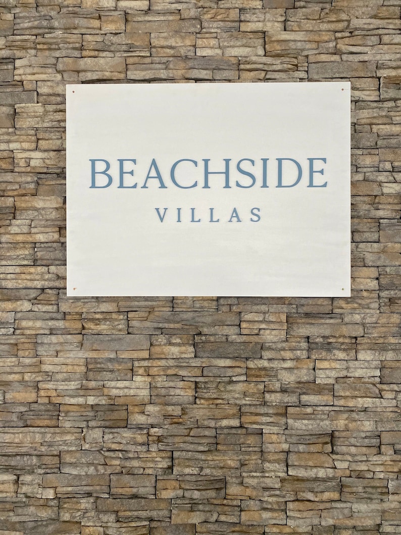 Puede incluir: Un cartel blanco con texto azul que dice "BEACHSIDE VILLAS" sobre un fondo de pared de piedra.