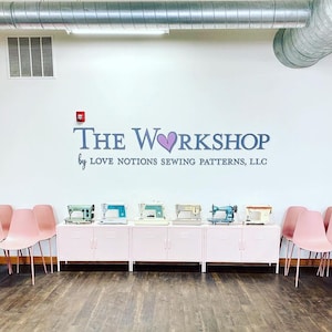 May include: A white wall features the text THE WORKSHOP by LOVE NOTIONS SEWING PATTERNS, LLC with a purple heart icon. Below, six vintage sewing machines in various colors sit on a long, light pink metal cabinet flanked by rows of pink chairs.