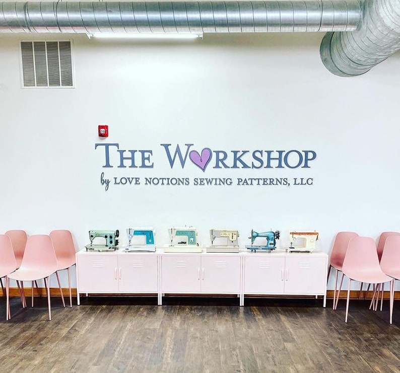 May include: A white wall features the text THE WORKSHOP by LOVE NOTIONS SEWING PATTERNS, LLC with a purple heart icon. Below, six vintage sewing machines in various colors sit on a long, light pink metal cabinet flanked by rows of pink chairs.