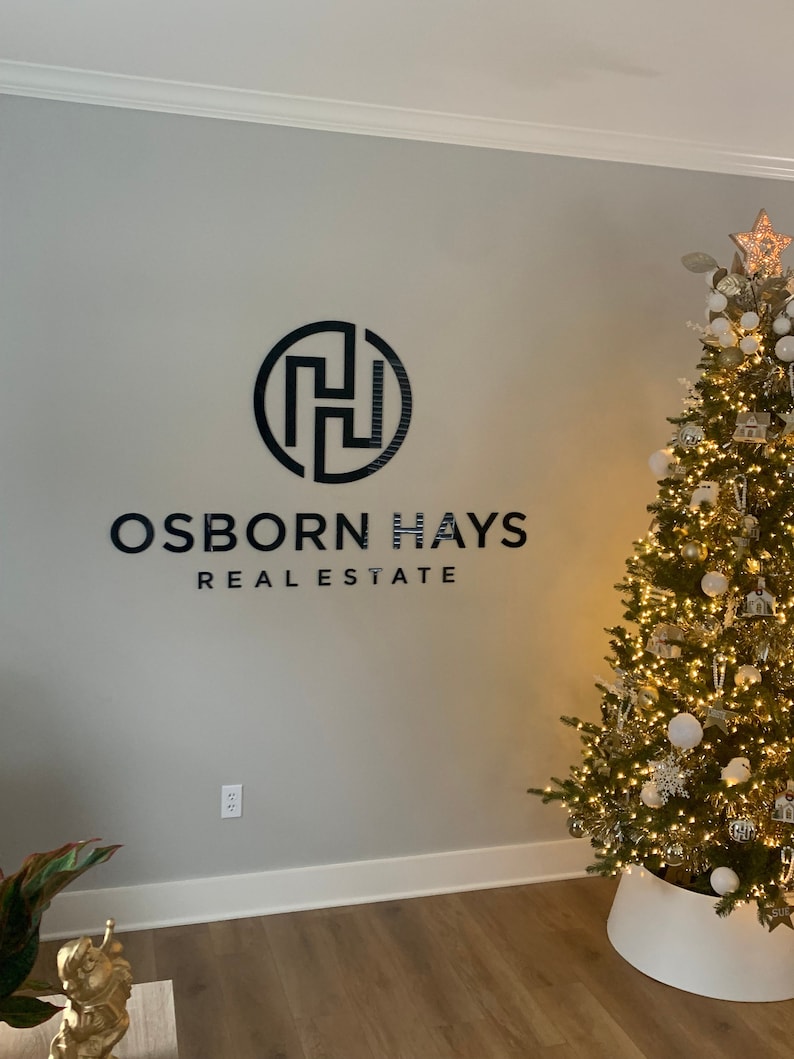 May include: A black wall-mounted sign featuring the text OSBORN HAYS REAL ESTATE and a circular logo with an abstract H design. The sign is positioned on a light gray wall next to a tall, illuminated Christmas tree decorated with white and gold ornaments.