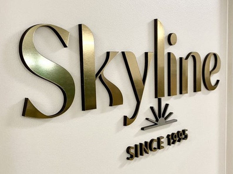 May include: A three-dimensional wall sign featuring the word Skyline in metallic gold lettering with black edges. Below the word is a small sun icon and the text SINCE 1995 in matching gold and black. The letters are mounted to a plain white surface.