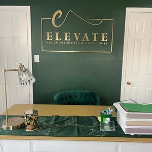 May include: A white desk with a gold top features a green runner, a gold lamp, a pineapple candle, and a document tray. On the dark green wall behind the desk, a gold sign reads ELEVATE MEDICAL AESTHETICS & WELLNESS CENTER with a stylized letter e.