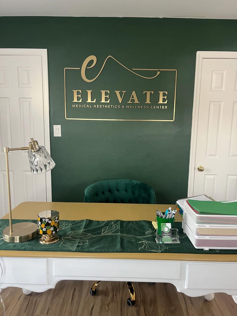 May include: A white desk with a gold top features a green runner, a gold lamp, a pineapple candle, and a document tray. On the dark green wall behind the desk, a gold sign reads ELEVATE MEDICAL AESTHETICS & WELLNESS CENTER with a stylized letter e.